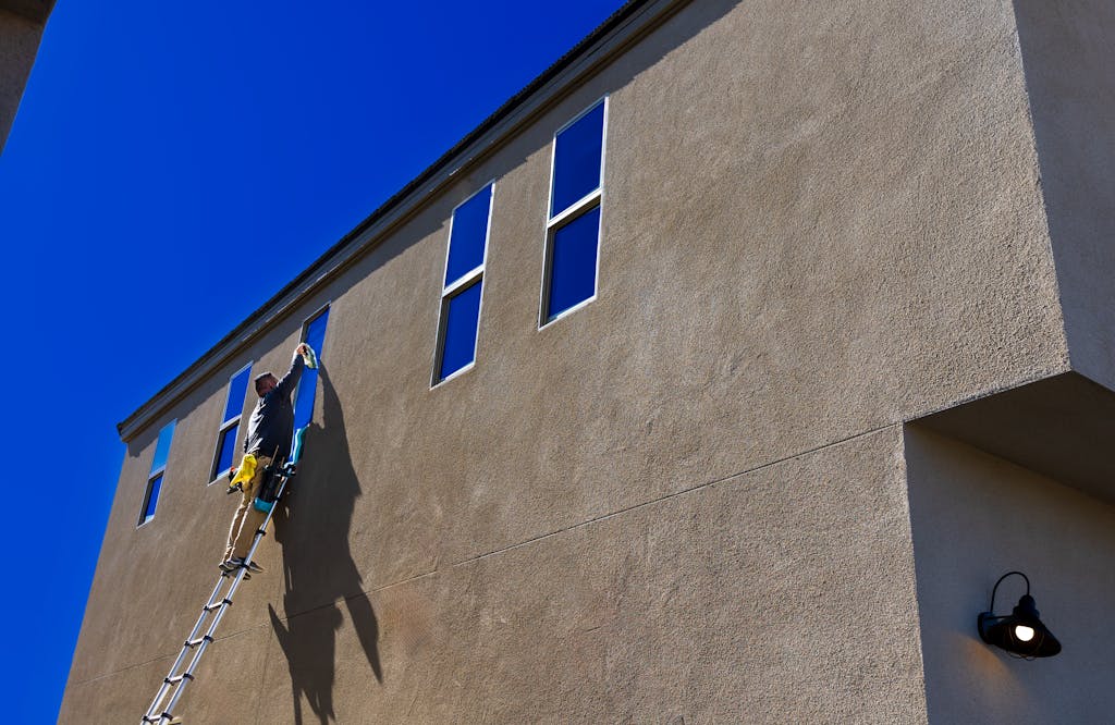 Man using a ladder to clean exterior windows of a tall building under a clear blue sky.