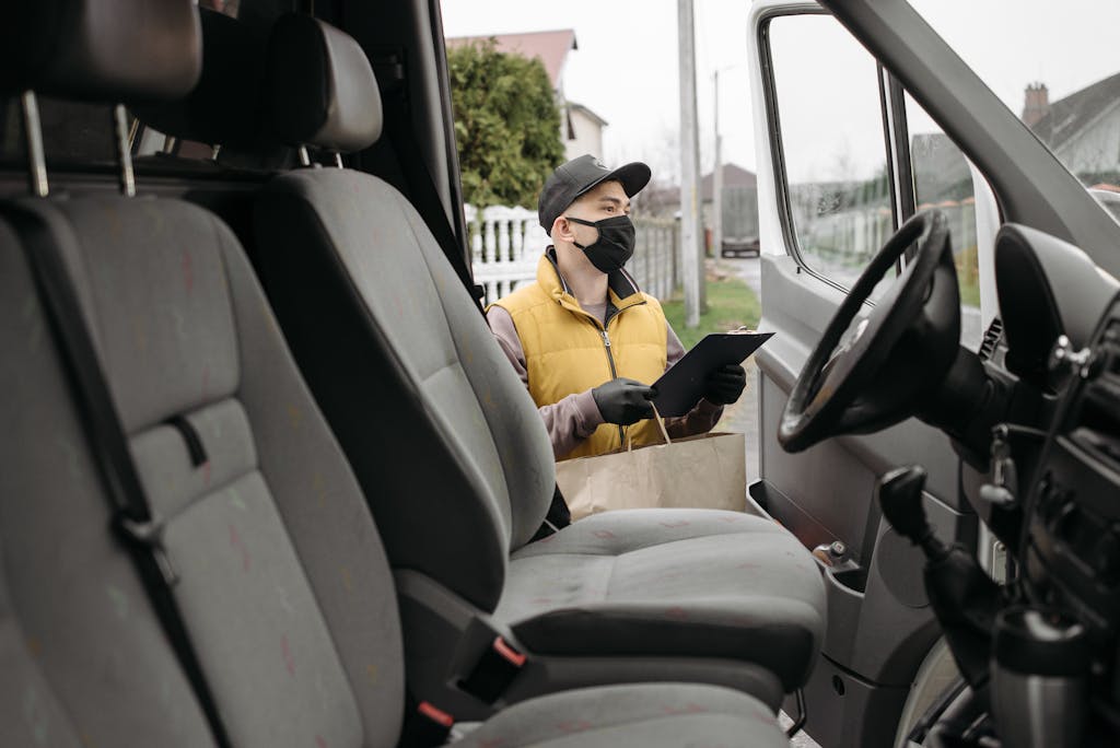 A delivery person wearing a mask stands outside a van holding a clipboard and bag, ready for home delivery.