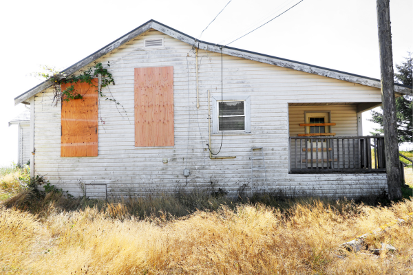 Old abandoned house with boarded-up windows, peeling white paint, and overgrown grass in front