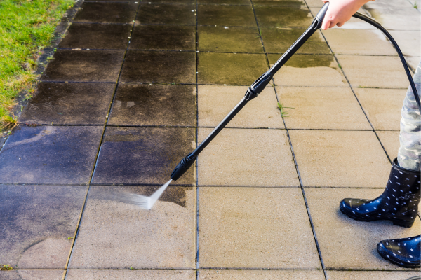 Person using a pressure washer to clean dirty pavement tiles, showing a clear before-and-after surface result.