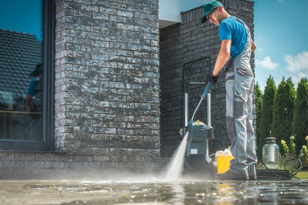 A worker is pressure-washing an outdoor surface beside a brick house, spraying away dirt with a high-powered hose.